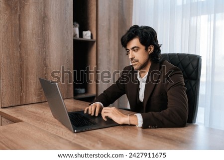 Handsome bearded businessman sitting at workplace by window, working on laptop, looking at screen, typing messages. Businessman dressed in suit concentrate working with notebook at workplace.