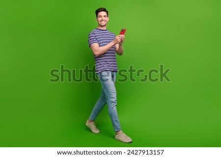 Full size photo of positive cool guy dressed striped t-shirt hold smartphone in arms go empty space isolated on green color background