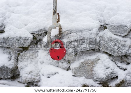 a heart-shaped red lock hanging at the background of grey stone wall covered with snow 