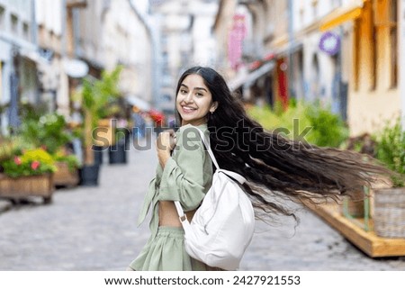 Photo of happy smiling Indian young girl walking, running looking back down city street, holding backpack and wind blowing hair.