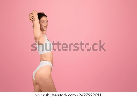 Side profile of a smiling caucasian woman in white sportswear stretching her arm behind her head, exuding fitness and joy on a pastel pink background. Wellness result, body care lifestyle Royalty-Free Stock Photo #2427929611