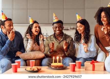 Joyful black student guy making birthday wish during party celebration with multiethnic friends, sitting together in living room interior, wearing festive hats. Bday celebration, friendship concept