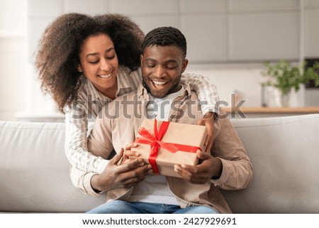 Young joyful black couple celebrating holidays and special occasions, woman presenting wrapped gift box to husband, sharing joy sitting on couch at home interior. Valentine's day present