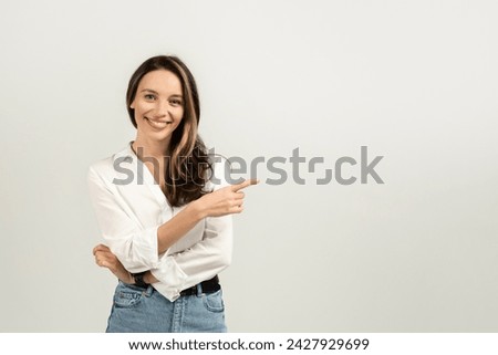 Confident and smiling european young brunette woman in a white blouse pointing to the side while standing against a plain light background with arms crossed, studio. Work, business ad and offer