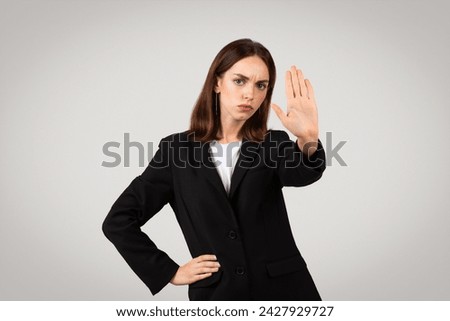 Stern millennial european businesswoman showing a stop hand sign with a serious expression, emphasizing boundaries or denial, in a formal black suit on a neutral background