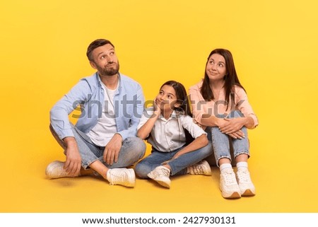 Family of three sitting together with pensive expressions, thinking and dreaming about something, posing over yellow studio background, shot of thoughtful parents and daughter girl Royalty-Free Stock Photo #2427930131