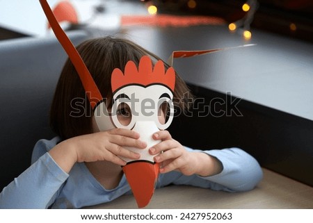 A child at a table in a children's cafe with a funny bird mask with a big beak near his face.                                Royalty-Free Stock Photo #2427952063