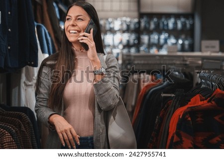 Beautiful young smiling woman walking in the retail store, looking at displayed clothes and having a joyful cell phone conversation.