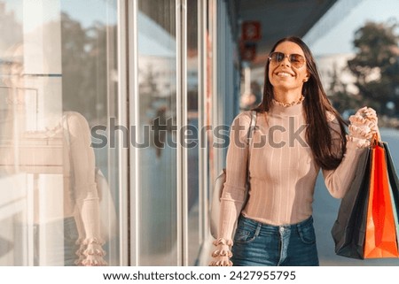 Young attractive woman holding colorful paper bags and watching shop windows while walking down the city streets during the sunny day.