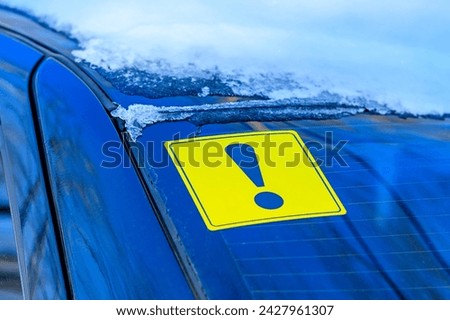 Yellow exclamation mark in selective focus on the rear window of a car with snow in winter. Designation of an inexperienced driver behind the wheel, a warning signal. Royalty-Free Stock Photo #2427961307