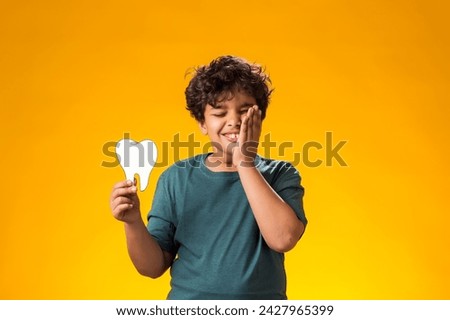 Portrait of child boy holding papercraft tooth, touching cheek because of toothache , over yellow background. Dental health concept