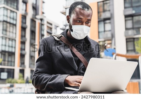 30s african man in mask against covid19 using his laptop in urban exterior. Businessman ceo manager freelancer typing working remotely while protecting against coronavirus spread