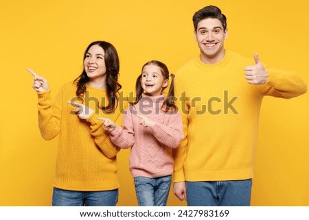 Young parents mom dad with child kid girl 7-8 years old wear pink sweater casual clothes point index finger aside on empty area show thumb up isolated on plain yellow background. Family day concept
