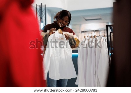 Clothing store buyer trying on shirt while exploring formal apparel for wardrobe. African american woman holding outfit on hanger, examining size, style and fit while shopping in boutique
