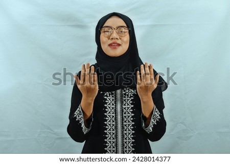 beautiful young Asian Muslim woman wearing hijab, glasses and black dress with white pattern raising hands and praying to god isolated on white background