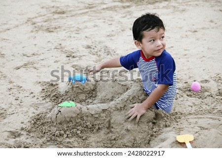 Exterior photo visual view of a cute funny eurasian asian kid child children boy who is playing at the beach in teh sand that covers his legs and he has lots of fun doing so using plastic shovel toys