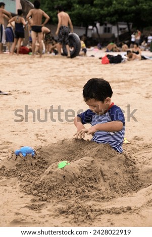 Exterior photo visual view of a cute funny eurasian asian kid child children boy who is playing at the beach in teh sand that covers his legs and he has lots of fun doing so using plastic shovel toys