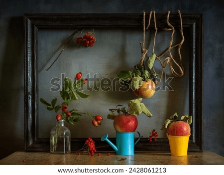 A dried red flower caught on an empty picture frame. On the table is a bottle with a rosehip branch, a watering can, a small bucket, apples with leaves.