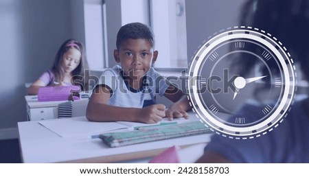 Image of clock over smiling biracial boy sitting at desk in diverse class. School, education, childhood and learning, digitally generated image.