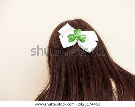 White bow with green shamrock attached on long brown hair of a girl. Light color background. Dressing up for special Irish community day. Popular symbol in Ireland. Celebrating Saint Patrick day