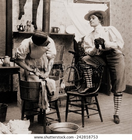 Humorous scene of life with 'the new woman.' Woman smoking and wearing knickers postures arrogantly as a man drudges over laundry. 1901. Royalty-Free Stock Photo #242818753