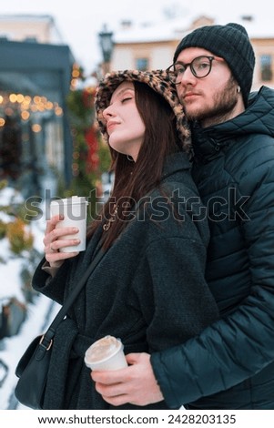 Stylish young beautiful couple of lovers in fashion winter clothes drink coffee, hug, rest and walks in the city on a winter day with snow
