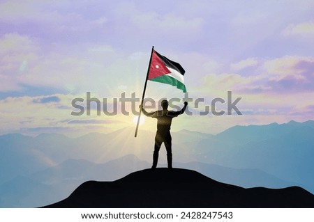Jordan flag being waved by a man celebrating success at the top of a mountain against sunset or sunrise. Jordan flag for Independence Day. Royalty-Free Stock Photo #2428247543