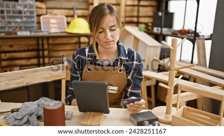 Attractive young blonde female carpenter, engrossed in serious business of financing her carpentry workshop, using credit card  touchpad amidst timber, indoor