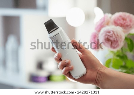 Ultrasonic skin scrubber in female hand, close up, mirror and flowers on the background. Concept of body and skin care, professional device