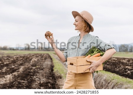 Female farmer holding wooden box full of fresh raw vegetables in agricultural field area. Woman standing proud with freshly bunch harvest outdoors. Local organic products, sustainable agriculture.