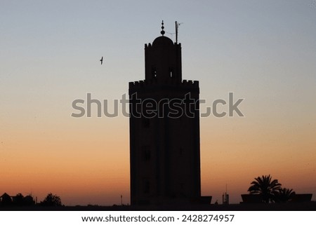 Portrait of scenic black silhouette of Moroccan minaret tower of old islamic mosque, palm trees and flying birds at beautiful orange sunset horizon sky in arabic oriental Marrakech, Morocco, Africa. Royalty-Free Stock Photo #2428274957