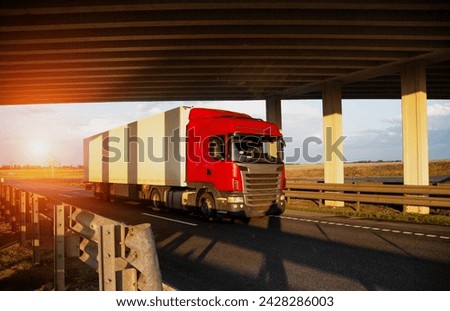 A red tractor with an isothermal refrigerated semi-trailer transports perishable goods with temperature conditions against the backdrop of sunset under the bridge. Delivery of perishable products  Royalty-Free Stock Photo #2428286003
