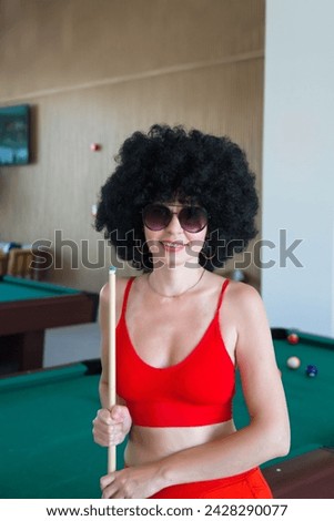 Girl who loves billiards. Woman with black curly hair in red clothes with glasses in the billiard room.