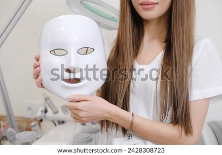 A cosmetologist in a professional setting holds a white LED face mask, possibly for skin rejuvenation treatments. Horizontal photo