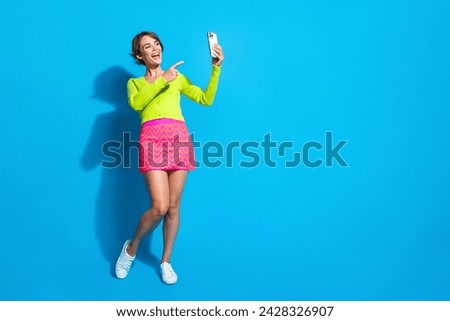 Full body length young positive lady point finger into smartphone display and talking with friends isolated on aquamarine color background