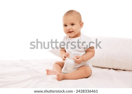 Little baby child, girl, adorable infant in white onesie sitting on bed with pillow and smiling against white background. Concept of childhood, family, care, motherhood, infancy, heath Royalty-Free Stock Photo #2428336461