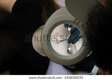 A cosmetologist during a facial cleansing session using special equipment to treat the client's skin, view through a magnifying lamp. Horizontal photo