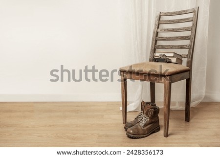 Brown leather hiking boots resting under a emaciated wooden chair with an old camera resting on the back along with some books Royalty-Free Stock Photo #2428356173