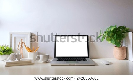 Laptop computer with blank display, potted plant, picture frame and stationery on white table.