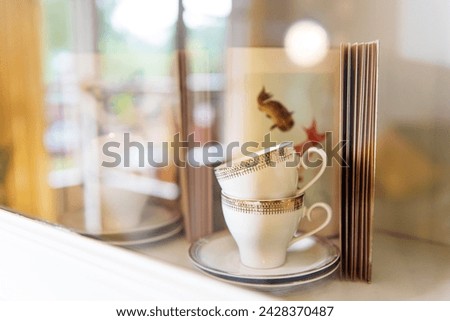 Shelf of grandmother’s china cabinet containing old book with goldfish illustration, stacked ceramic cups and saucers with gold embossing and other out-of-focus objects behind glass door Royalty-Free Stock Photo #2428370487