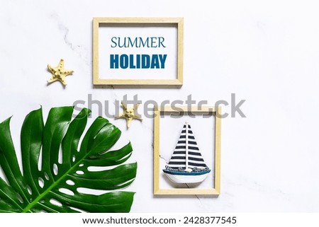 Summer vacation concept. Artistic composition with big tropical monstera leaf, decorative star fish and two wooden picture frames with a boat and text Summer Holiday on white marble background.