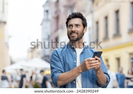A smiling young man walks down the city street in casual clothes and holds a mobile phone in his hands, looks for a place and meeting on the navigator, looks to the side. Close-up photo.