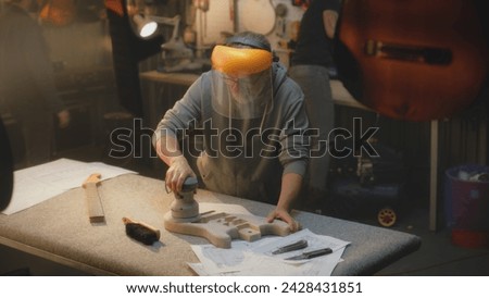 Caucasian mature artisan in protective mask grinds guitar body using grinding machine. Craftsman tries on guitar fretboard. Carpenter makes electro guitar. Concept of handcraft and small business. Royalty-Free Stock Photo #2428431851