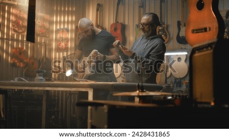 Two professional carpenters make wooden guitar and take measurements. Craftsmen in modern workshop for making musical instruments. Colleague works in the background. Handmade and entrepreneurship.