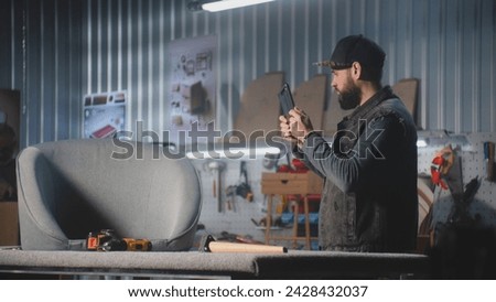 Male carpenter or designer takes photo of chair using tablet computer, works in stylish workshop for making furniture. Mature colleague assembles shelf in the background. Handcraft and small business.