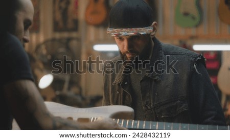 Close up of middle aged carpenters making electro guitar in stylish workshop. Professional tattooed artisan talks with colleague and tries on guitar fretboard. Handcraft and small business concept. Royalty-Free Stock Photo #2428432119
