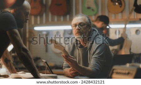 Male mature artisan uses digital tablet computer to draw blueprints of guitar. Middle aged tattooed carpenter talks with colleague, takes guitar fretboards. Craftsmen work in light stylish workshop. Royalty-Free Stock Photo #2428432121
