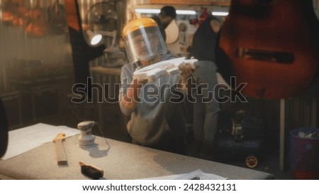 Caucasian mature artisan in protective mask grinds guitar body using grinding machine. Craftsman tries on guitar fretboard. Carpenter makes electro guitar. Concept of handcraft and small business. Royalty-Free Stock Photo #2428432131