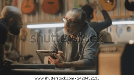 Male mature artisan uses digital tablet computer to draw blueprints of guitar. Middle aged tattooed carpenter talks with colleague, takes guitar fretboards. Craftsmen work in light stylish workshop. Royalty-Free Stock Photo #2428432135