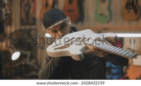 Close up of middle aged carpenters making electro guitar in stylish workshop. Professional tattooed artisan talks with colleague and tries on guitar fretboard. Handcraft and small business concept. Royalty-Free Stock Photo #2428432159